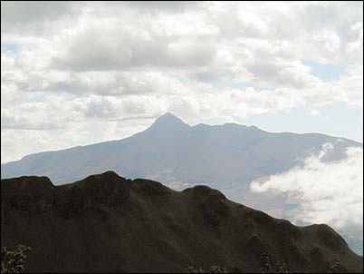 En primer plano se nota parte del cr�ter del Pasochoa desde donde se realiza esta toma, en segundo plano y de perfil el volc�n Coraz�n. La caldera del volc�n Coraz�n se ha erosionado fuertemente apareciendo ahora como una profunda quebrada. El estudio de los volcanes ecuatorianos nos dan una idea bastante cercana de como los grandes edificios volc�nicos se erosionan hasta hacerse irreconocibles.
